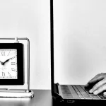 a person typing on a laptop next to a clock