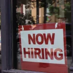 A "now hiring" sign hangs in a store window.