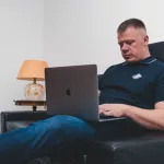man in black polo shirt sitting on black leather couch using macbook