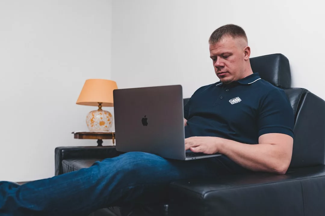 man in black polo shirt sitting on black leather couch using macbook